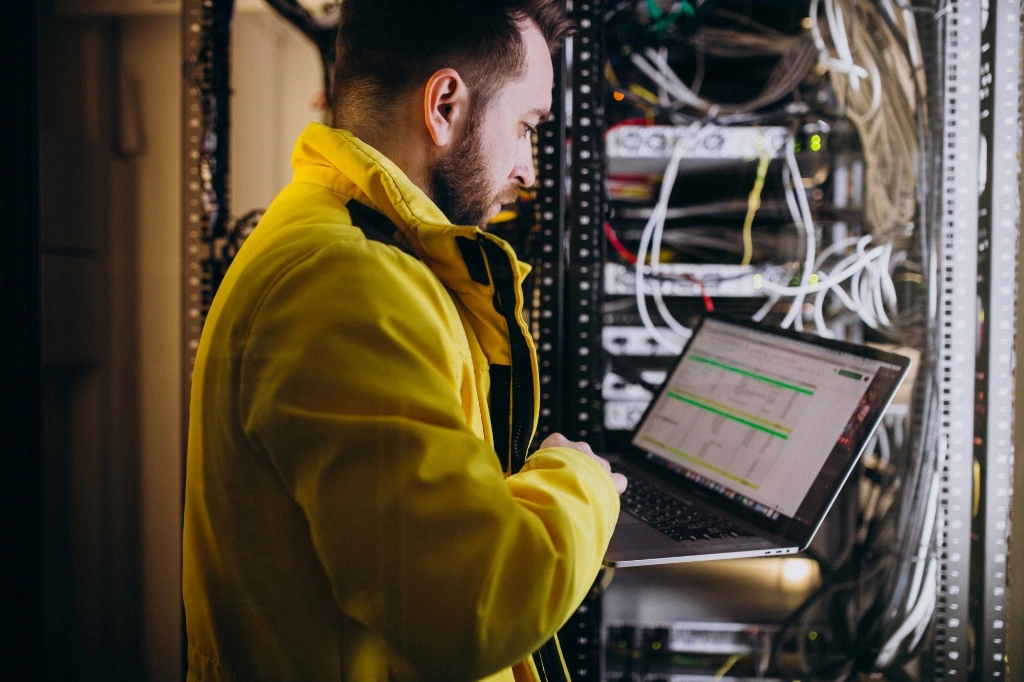 A technical engineer in a yellow jacket using a laptop to verify an FPGA schematic design while standing in front of a server rack in a data center.